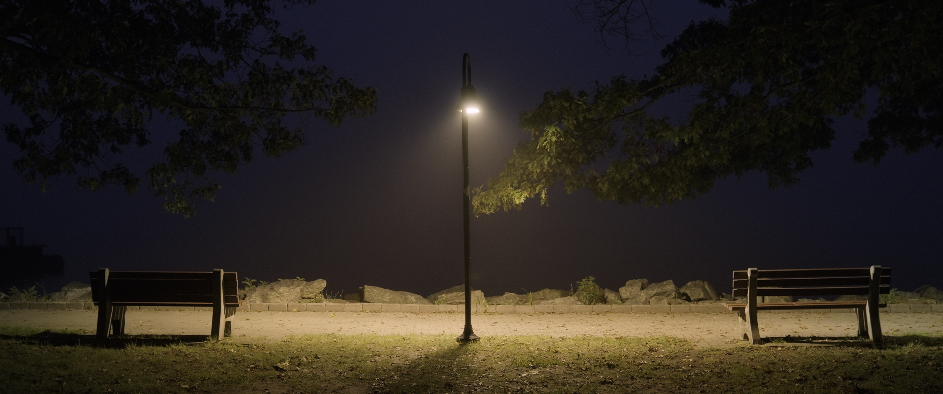 Warm lamplight between two empty benches beside the water at night